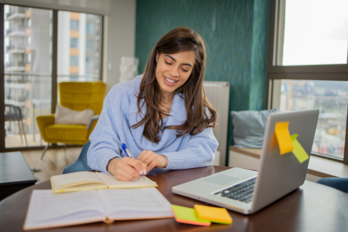 Professional sitting at a desk with their laptop. They are writing notes and smiling. 