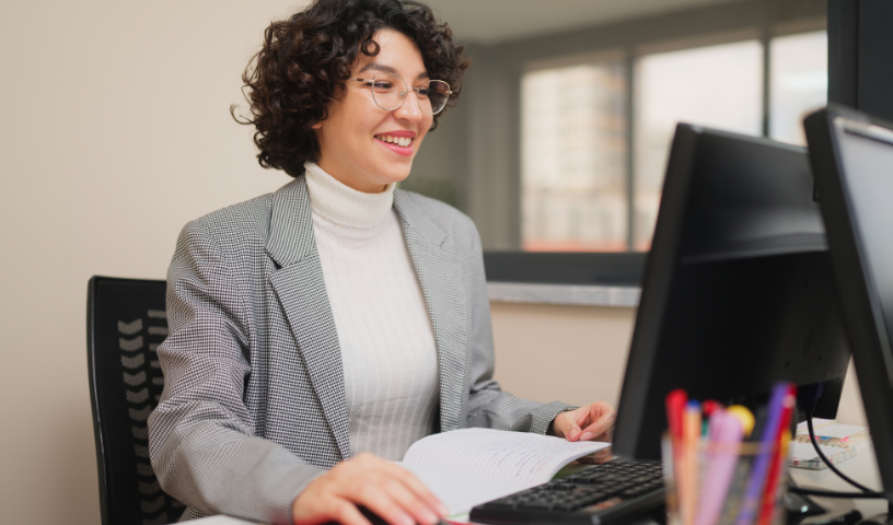 A professional sits at their desk, they are holing a mouse, looking towards the desktop screen and smiling. 