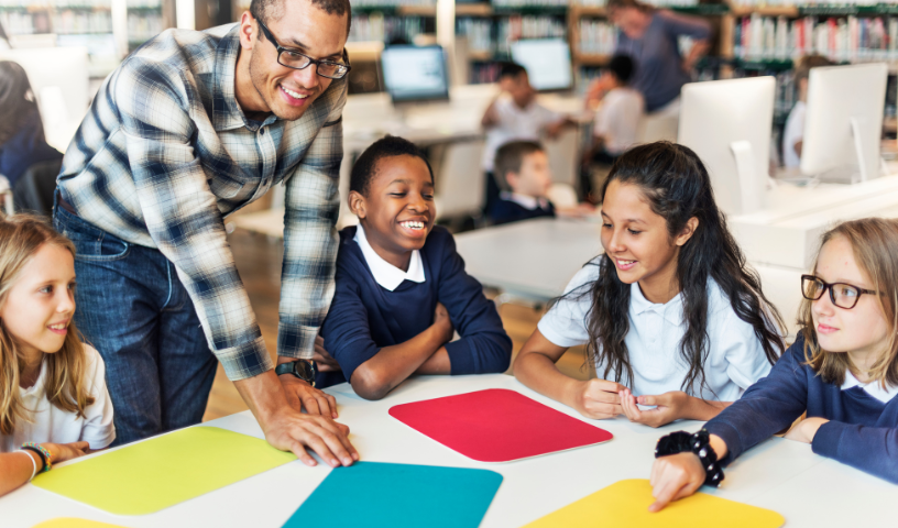 A group of primary children sit around a desk. A Teacher stands over them and talks. 