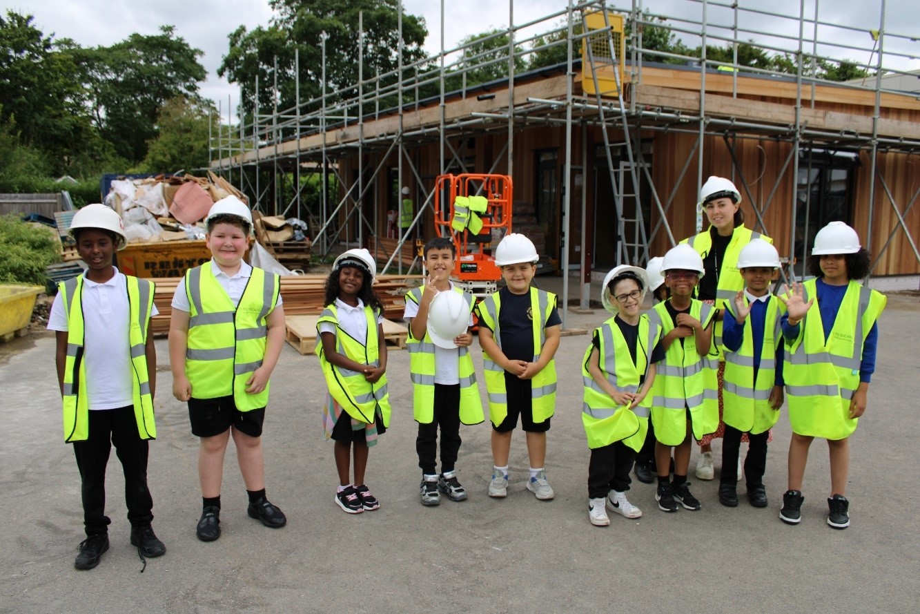 A group of children stand on a building site. They are all wearing high vis jackets and helmets