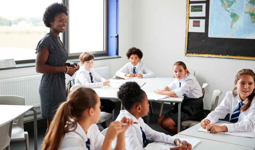 A teacher stands in a classroom with secondary school pupils sitting at their desks. The pupils are in white shirts and blue striped ties. 