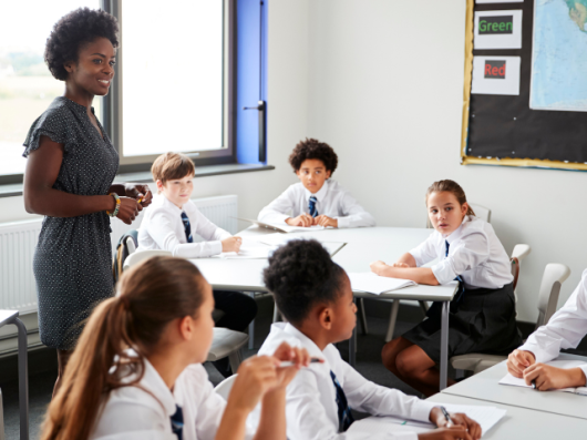 Teacher stands in classroom, a group of pupils sitting at their desks around them.