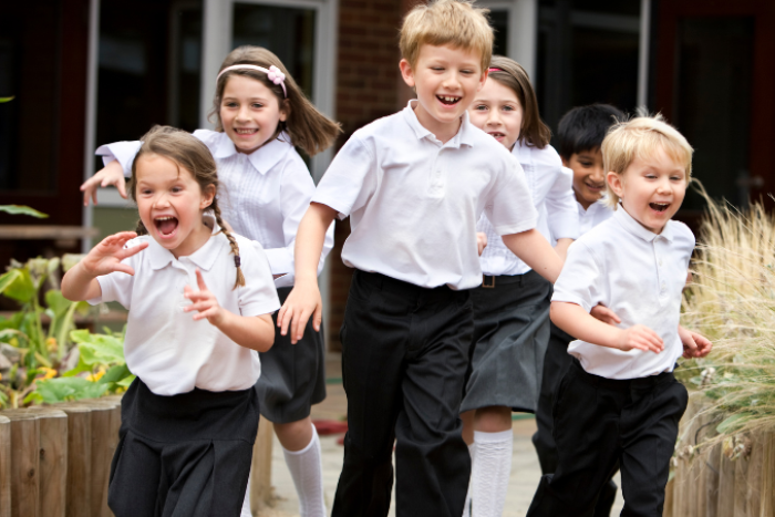 A group of primary school pupils in white polo shirts are running through a playground. They are all smiling and some of them cheering. 