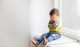 A toddler sits on a windowsill with their hands held over their face. 