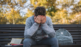 A young person sitting on a bench with their backpack and book sitting next to them. They have their head in their hands. 