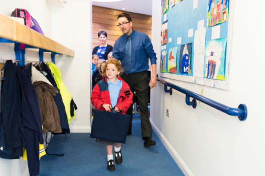 A primary aged pupil walks into the school corridor with their book bag and coat. A teacher is walking closely behind them. 