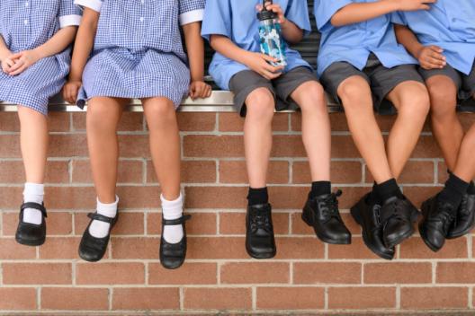 A group of primary school pupils sit on a wall in a row. Their legs dangle over the edge and you can see they shiny leather school shoes. 