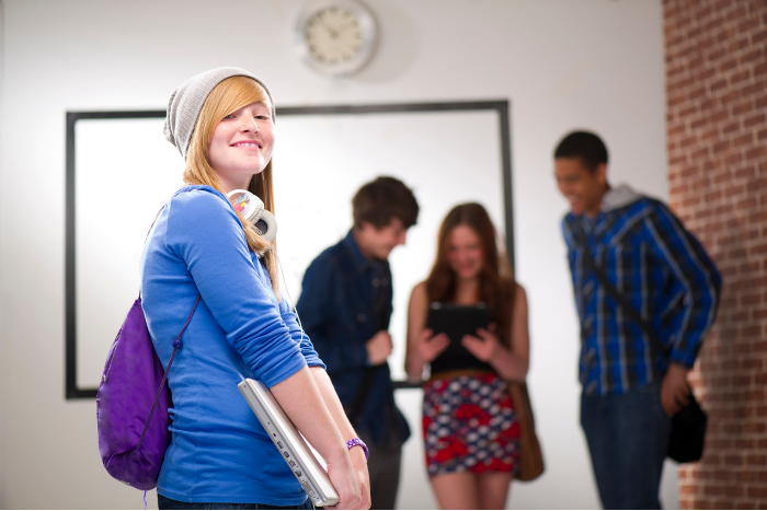 Sixth form student holding laptop, looking to the side at camera, smiling. 